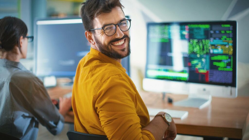 A web developer smiles and looks back at the camera while his desktop computer displays web development skills.