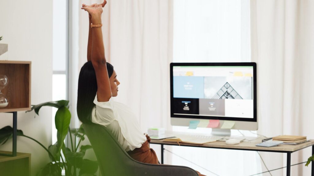 A woman stretches at her wooden desk, which has a desktop computer on it, demonstrating her WordPress expertise.