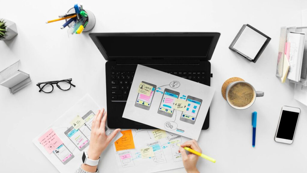 A black laptop is displayed on a white desk with pieces of paper over it, demonstrating web design mockups and possibilities.