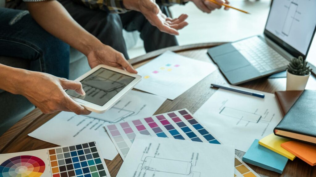 A laptop and tablet are displayed on a wooden desk. Two people sit in front of the desk and are looking at color swatches and more for web design purposes.