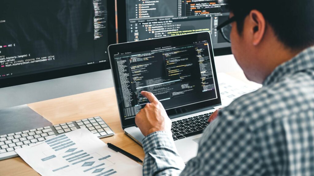 A web developer wearing a plaid shirt looks at a laptop that has web development solutions displayed. There is a desktop computer behind the laptop and a second monitor to the right.