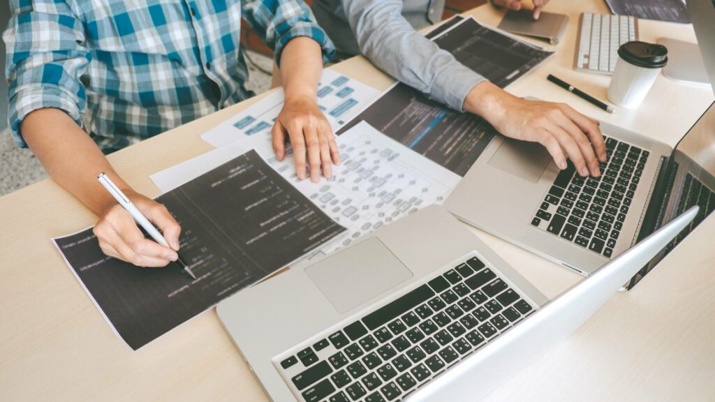 Two people sit at a white desk and work on two laptops with printed paper of development code displayed in front of them.