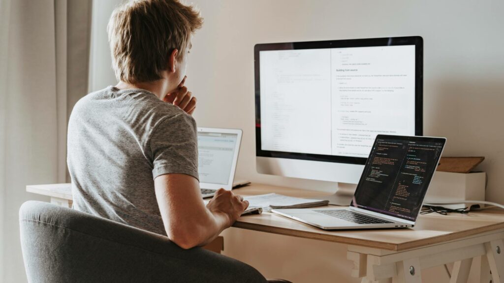 A person sits at a wooden desk and works on two laptops and a desktop, demonstrating front end development.