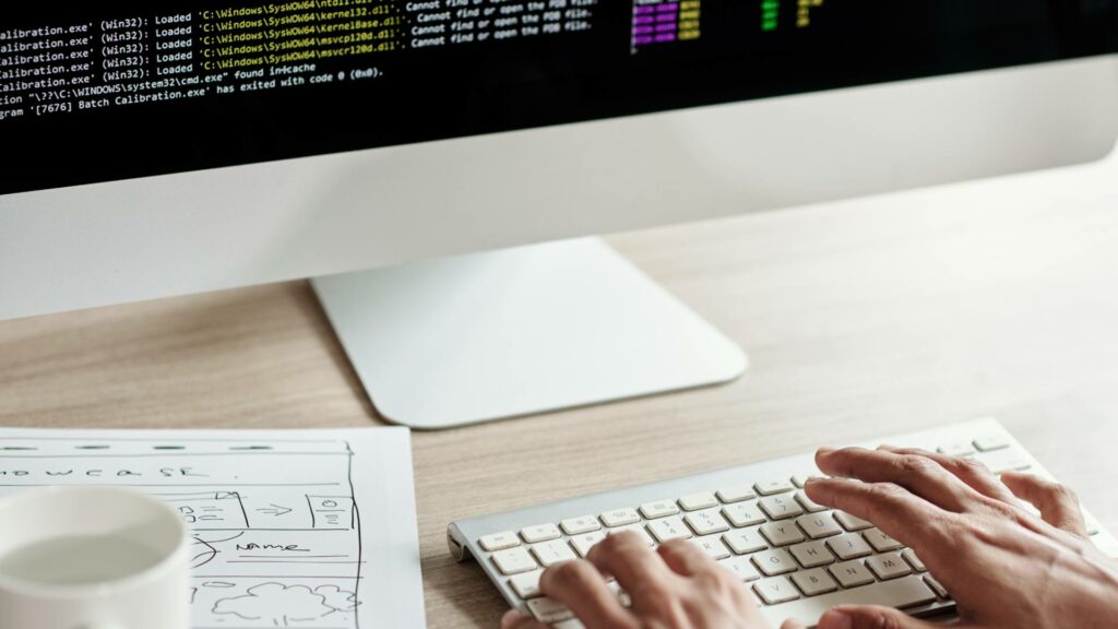 A person types on a keyboard while front end development code is displayed on a desktop computer.