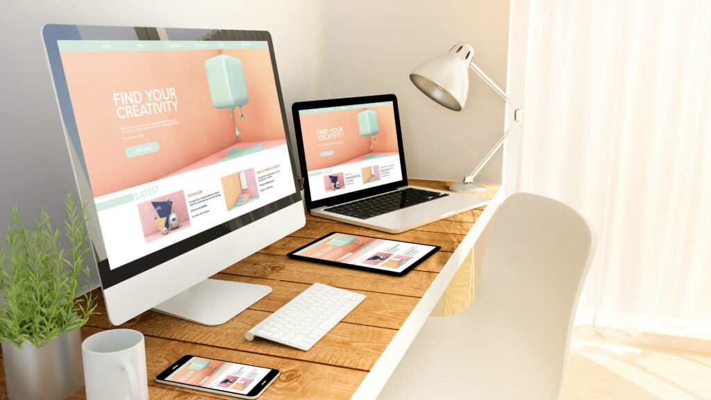 Responsive design is displayed on a desktop, laptop, tablet, and phone on a wooden desk.