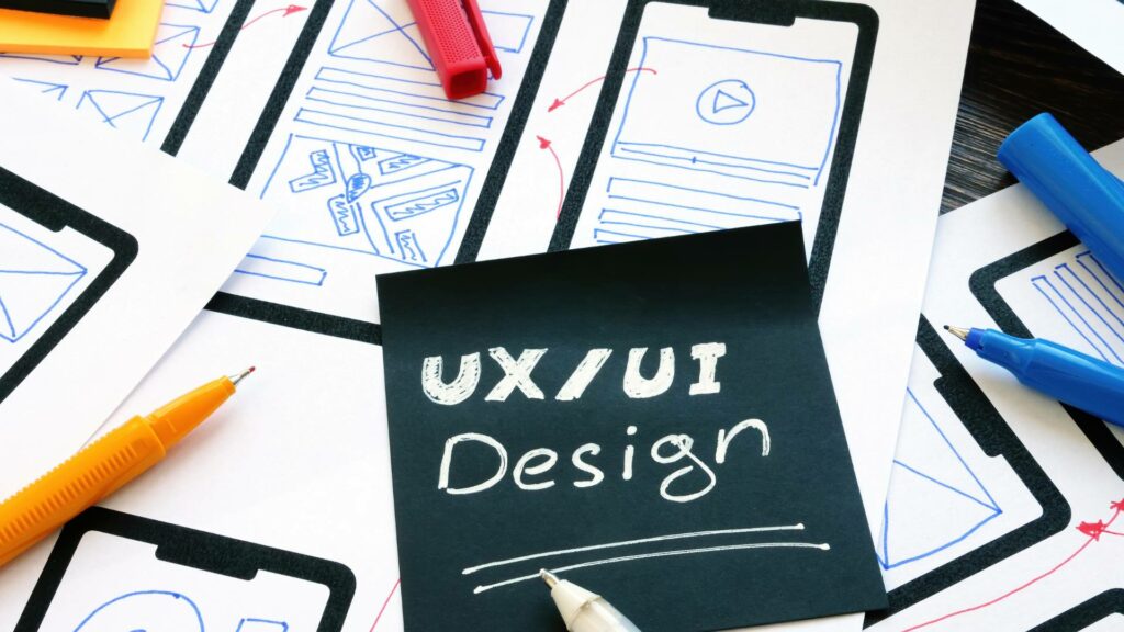 A board with the words UX UI Design are displayed atop papers with user experience friendly designs.