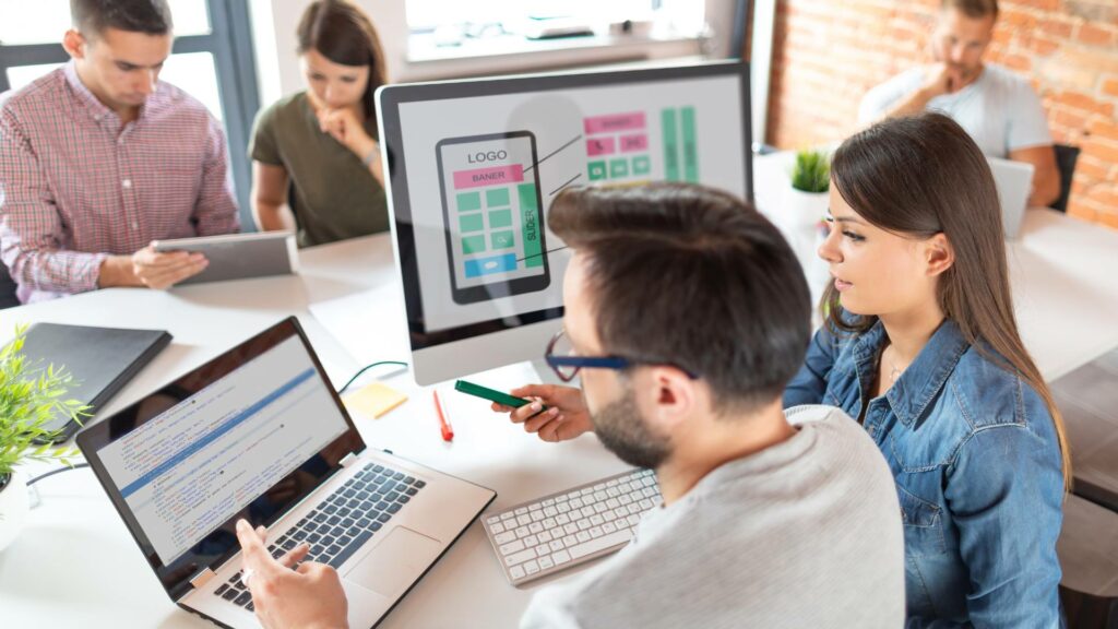 Two works sit at a large coworking table as they work on a desktop and laptop, demonstrating ux/ui design.