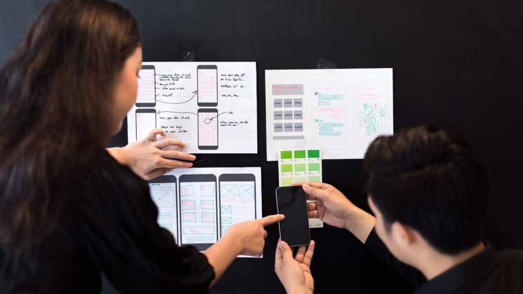 Two people work on UI design by holding up their cell phone and comparing it to displayed papers with design options on them.