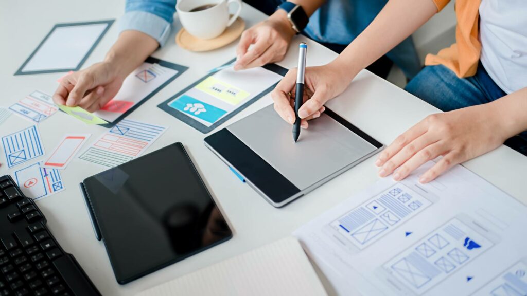 Two people are sitting at a white desk working on tablets and a computer, demonstrating web development and Wordpress skills.