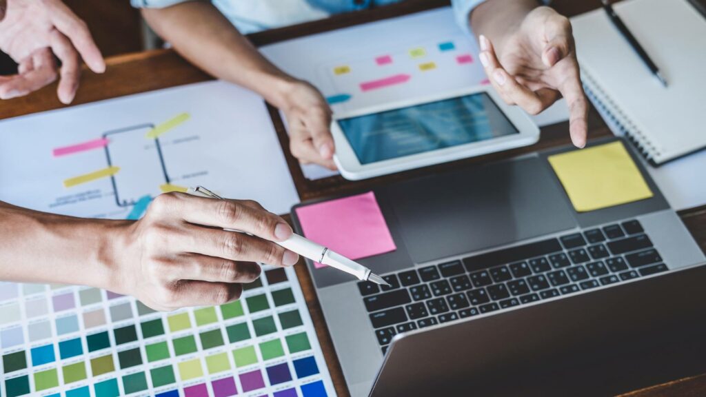 Two people work on a laptop and tablet, and are looking at color swatches laid out on the desk, demonstrating essential web development skills 2024.