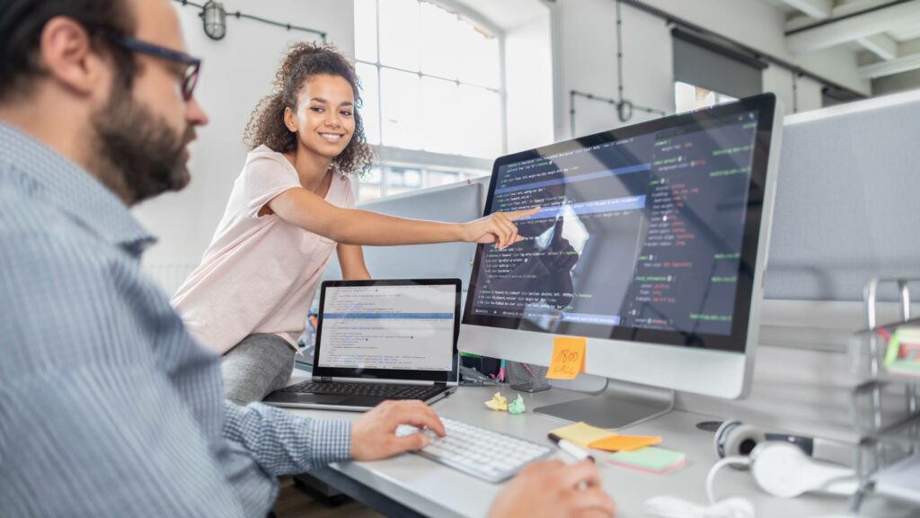 A man sits at a desk working on a desktop computer and laptop, while a woman to his left smiles and points at the desktop screen, demonstrating back end and front end code and solutions.