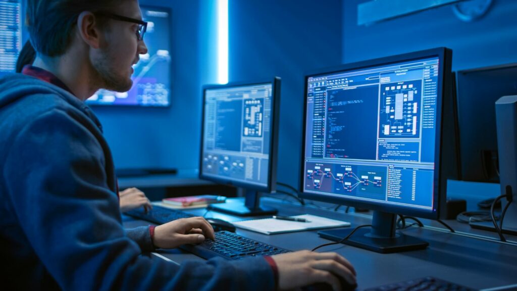 A person sits in a room that has blue lighting and works on two desktop computers, demonstrating back end systems and development.