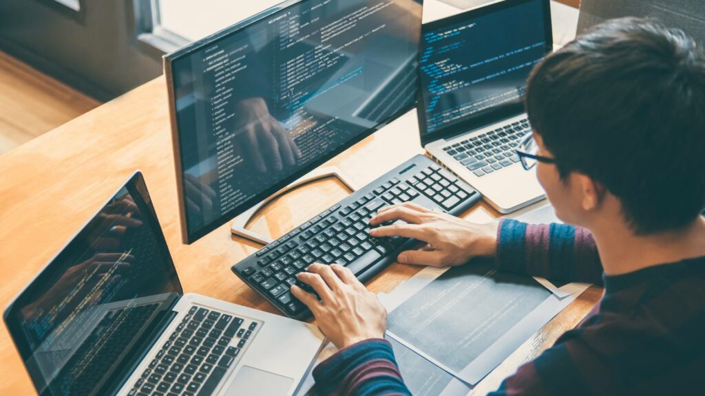 A person sits at a wooden desk and works on one desktop and two laptop, demonstrating back end systems and development.