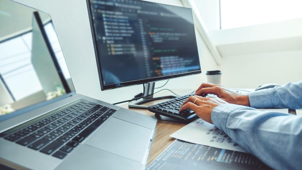 A person types on a desktop keyboard and there's another laptop to their left. They are working on software development code on a dark screen.