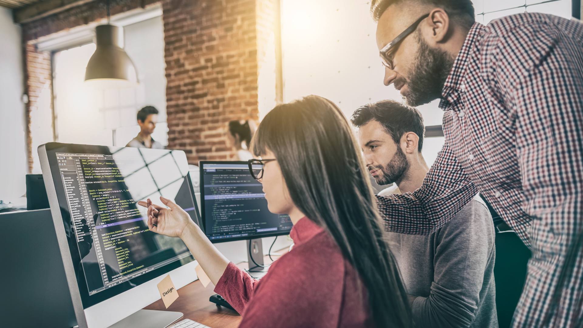 Three people sit around a desktop computer and figure out software development code.