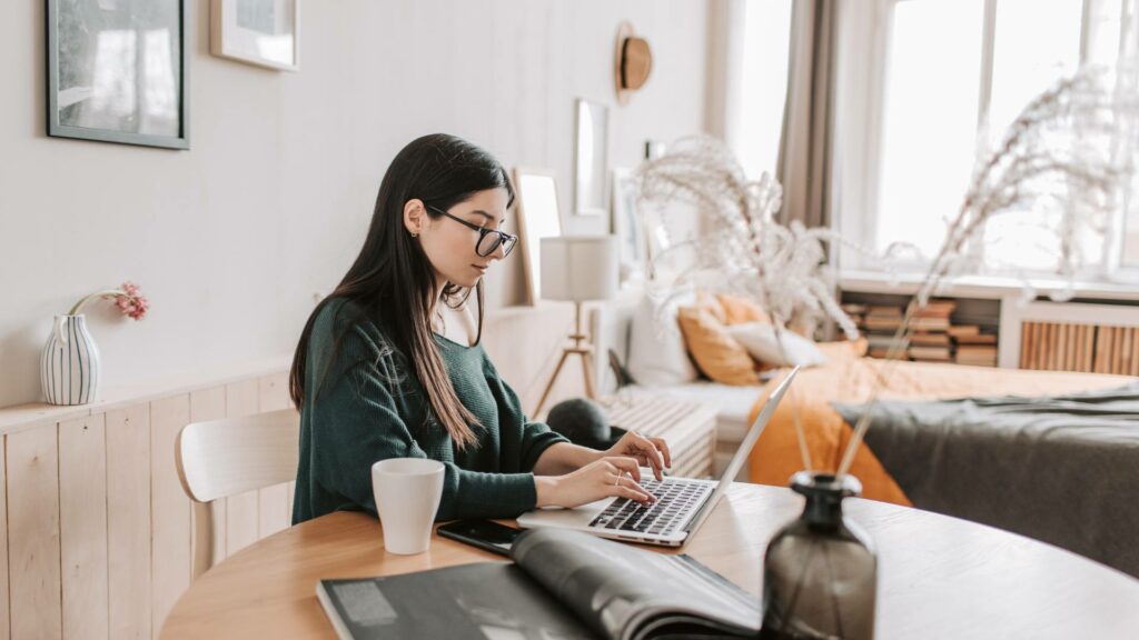 A person sits at a wooden table at writes on her laptop, demonstrating writing a newsletter in a studio apartment.