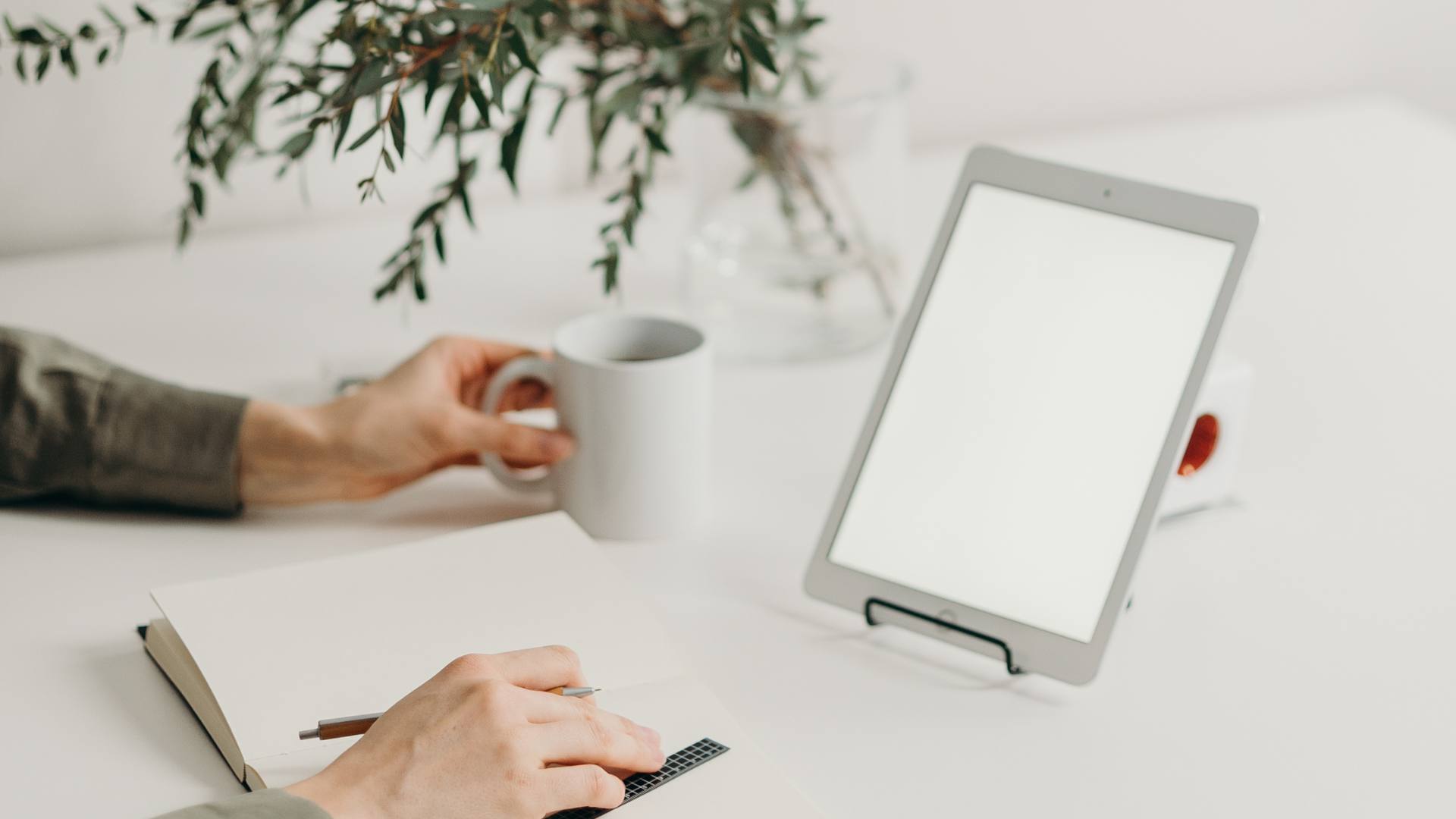 A person sits at a table with a white mug and writes in their notebook as their tablet is up, demonstrating a newsletter.